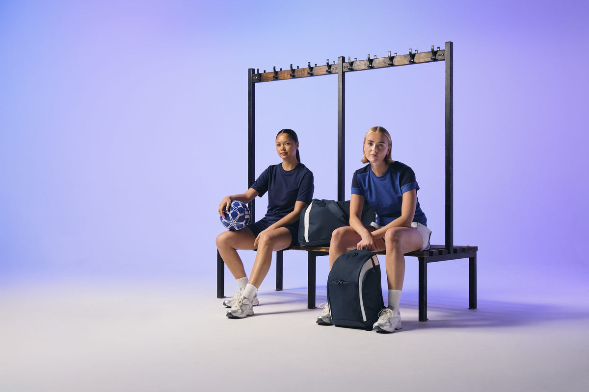 Two footballers on a changing-room bench with BagBase navy kit bags