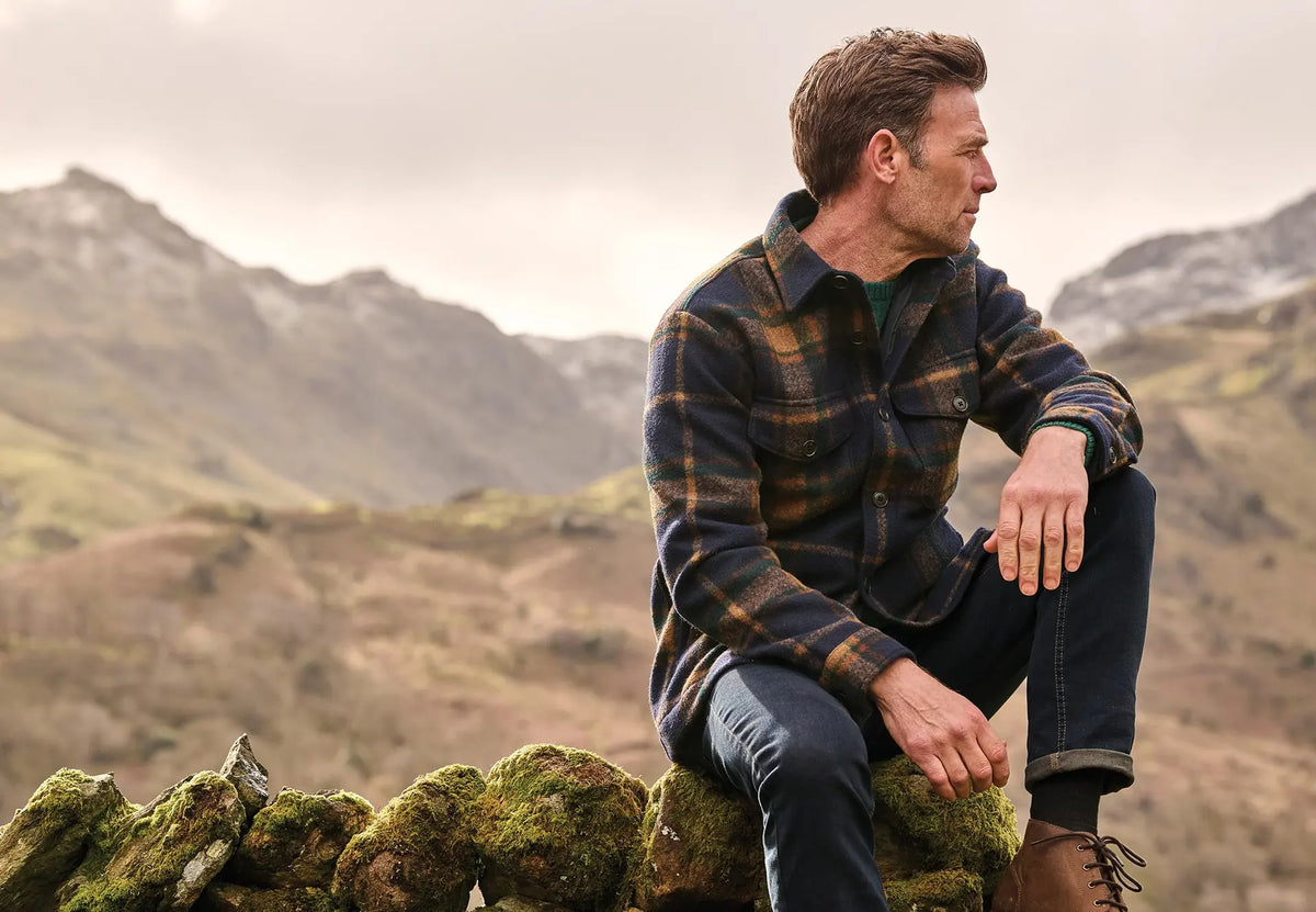 Man in a Brook Taverner check overshirt sitting on a stone wall in the hills